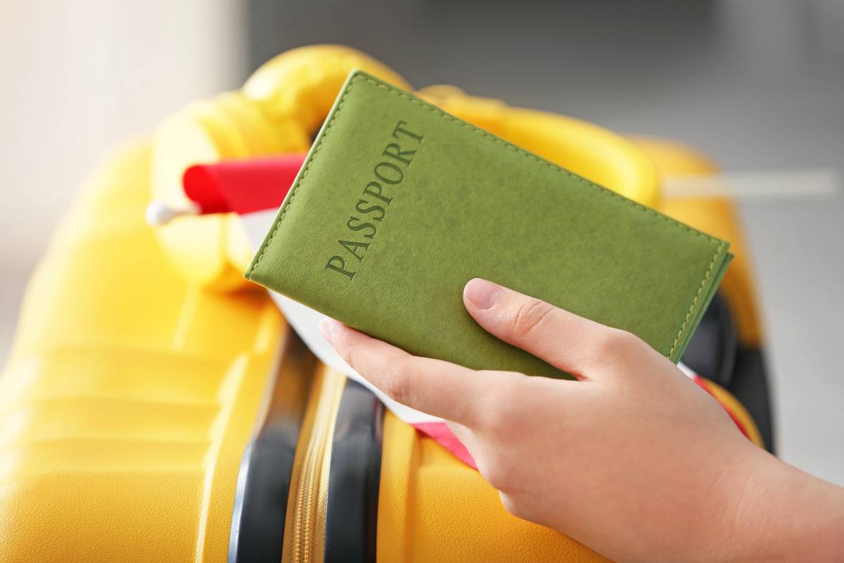 Une main tient un étui vert marqué Passport devant une valise jaune.